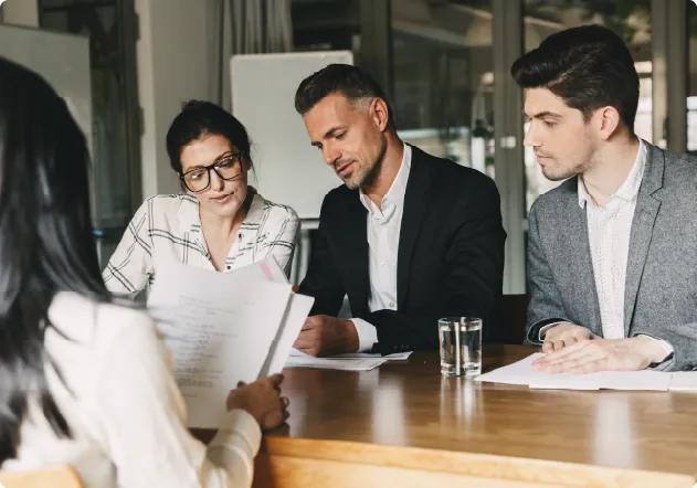 three people around a desk looking at a document and someone sat opposite looking at a document